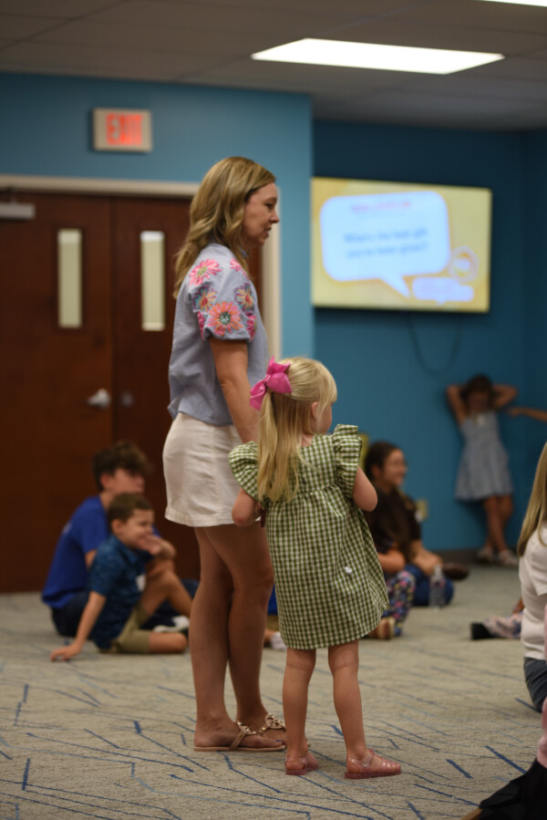 A Kids Ministry leader standing with a young girl in the kids room, with other children sitting on the floor nearby.