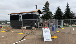 A tiny home with navy blue siding surrounded by chain link fence. Being built in the Valley Creek Campus parking lot.