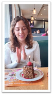 young hispanic woman claps her hands while looking at a chocolate slice of cake with a lit candle on it