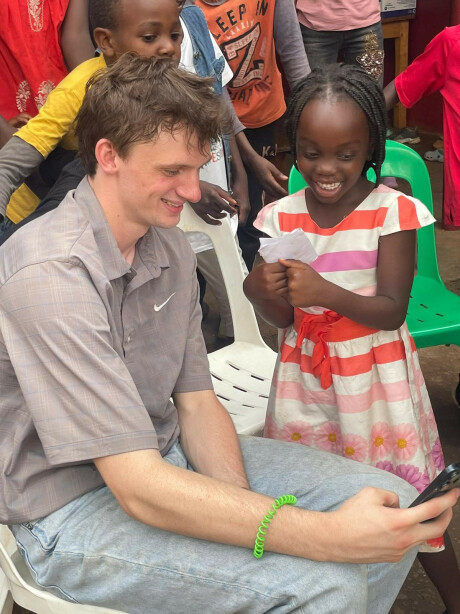 young man from FPC mission trip team wearing a grey shirt and blue jeans on his phone while a Ugandan boy in a yellow shirt and girl in a striped white orange and pink dress look over his shoulder at the screen