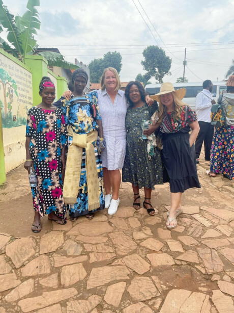 three Ugandan women in colorful floral dresses smiling and posing with two women from the FPC mission team on a cobblestone street