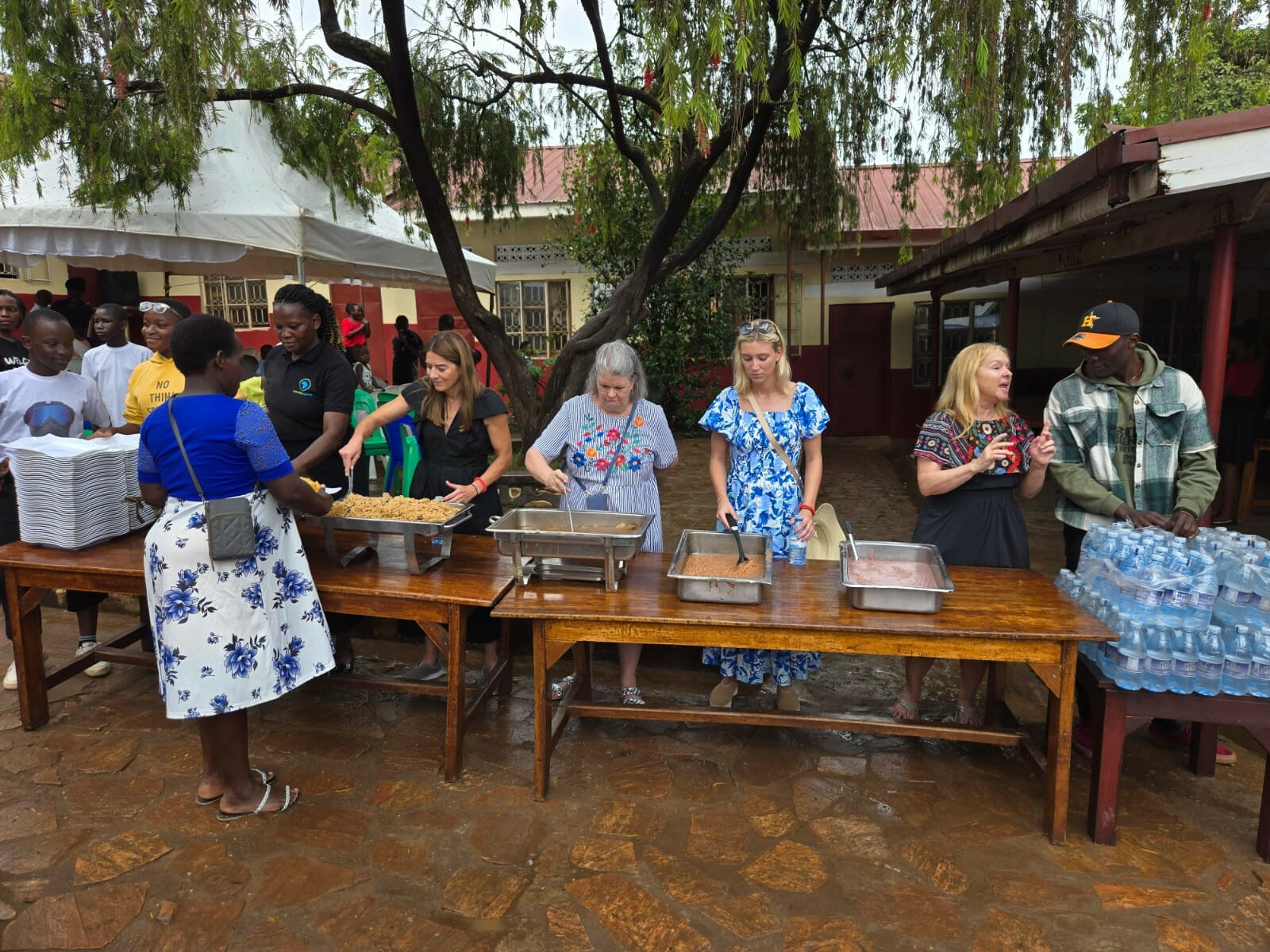 line of people at a lunch buffet on wooden tables under a large tree in an outdoor courtyard in Uganda