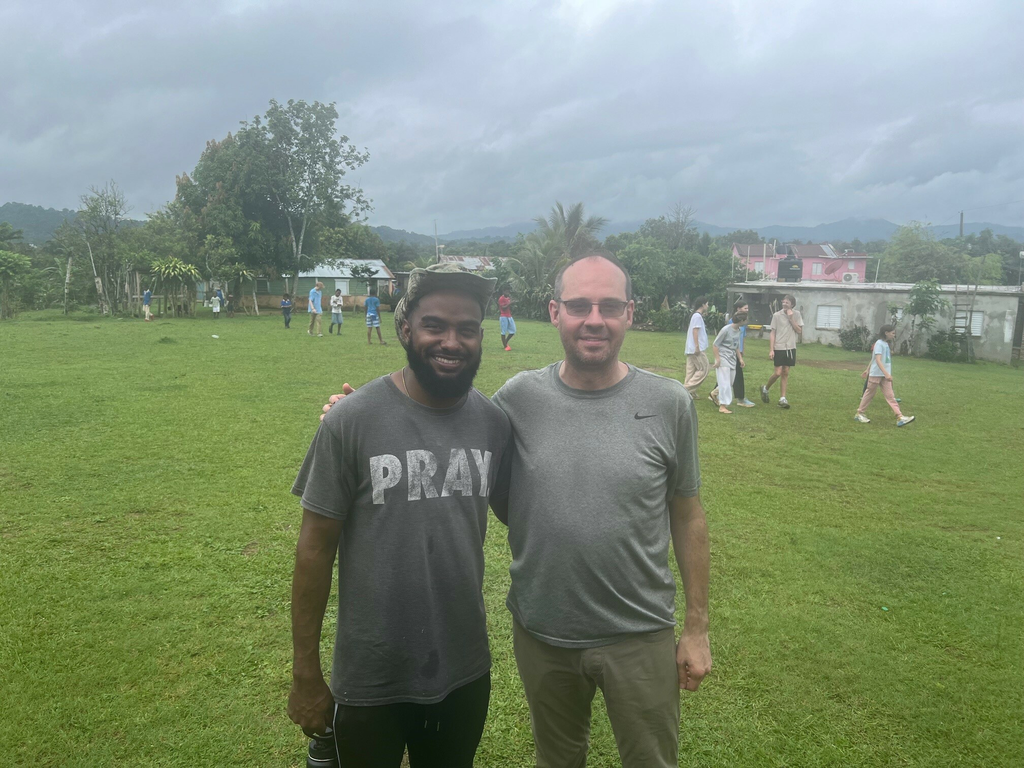 two men, one american and one dominican, standing in a field with children and houses in the background