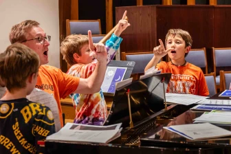 Children surrounding a grand piano work on their vocal technique
