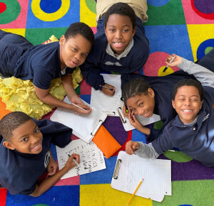 Logos Academy students smiling at the camera while laying on a colorful carpet and writing on clipboards