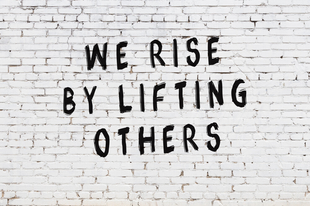 we-rise-by-lifting-others-quote-black-lettering-white-brick-wall